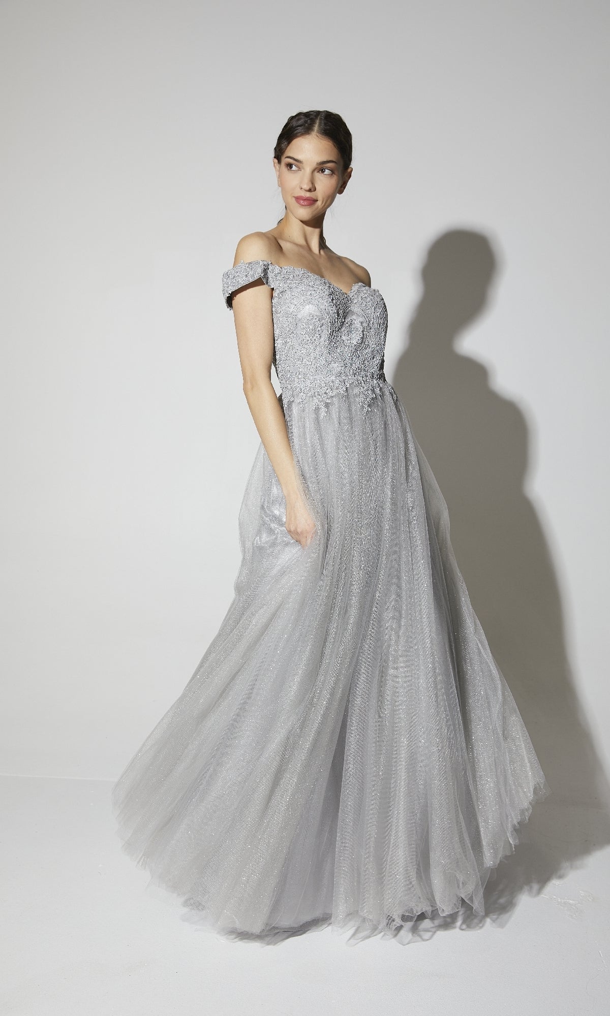A woman poses in the Off-Shoulder Glitter Prom Ball Gown: Eureka 9899, featuring a light gray, off-the-shoulder design with lace details and a tulle skirt, set against a plain white background.